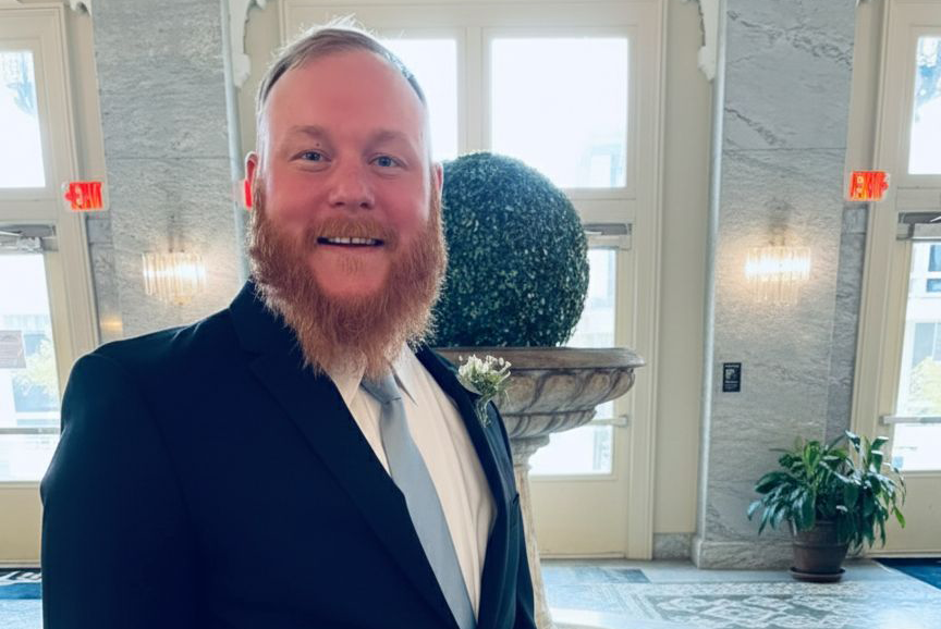 A man with a beard dressed in a suit smiles at the camera]