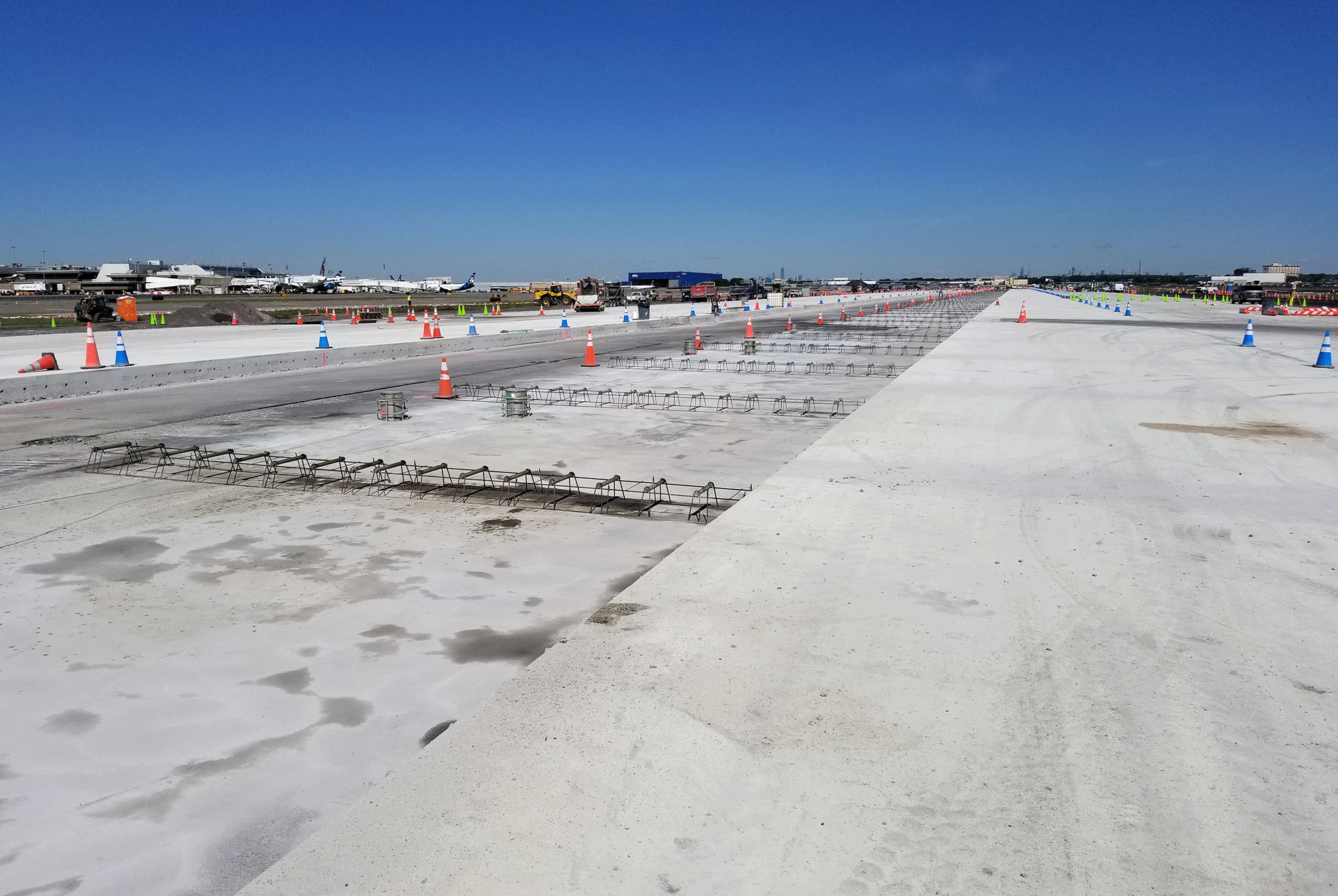 Airport runway under construction, with a long stretch of concrete, blue sky above, and traffic cones lining the path. Machinery and buildings in the distance.