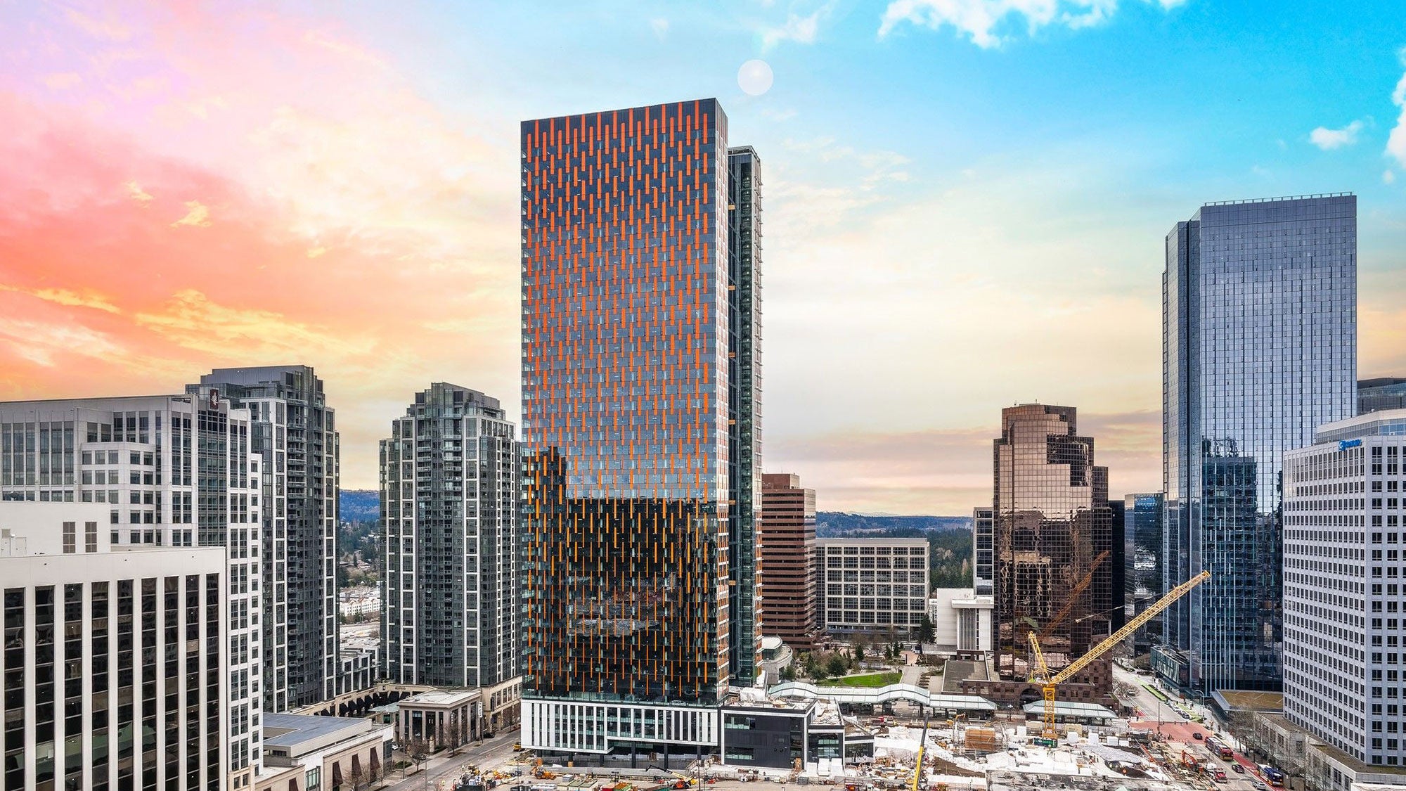 A reflective, high-rise building surrounded by other tall buildings with the sun setting behind it.