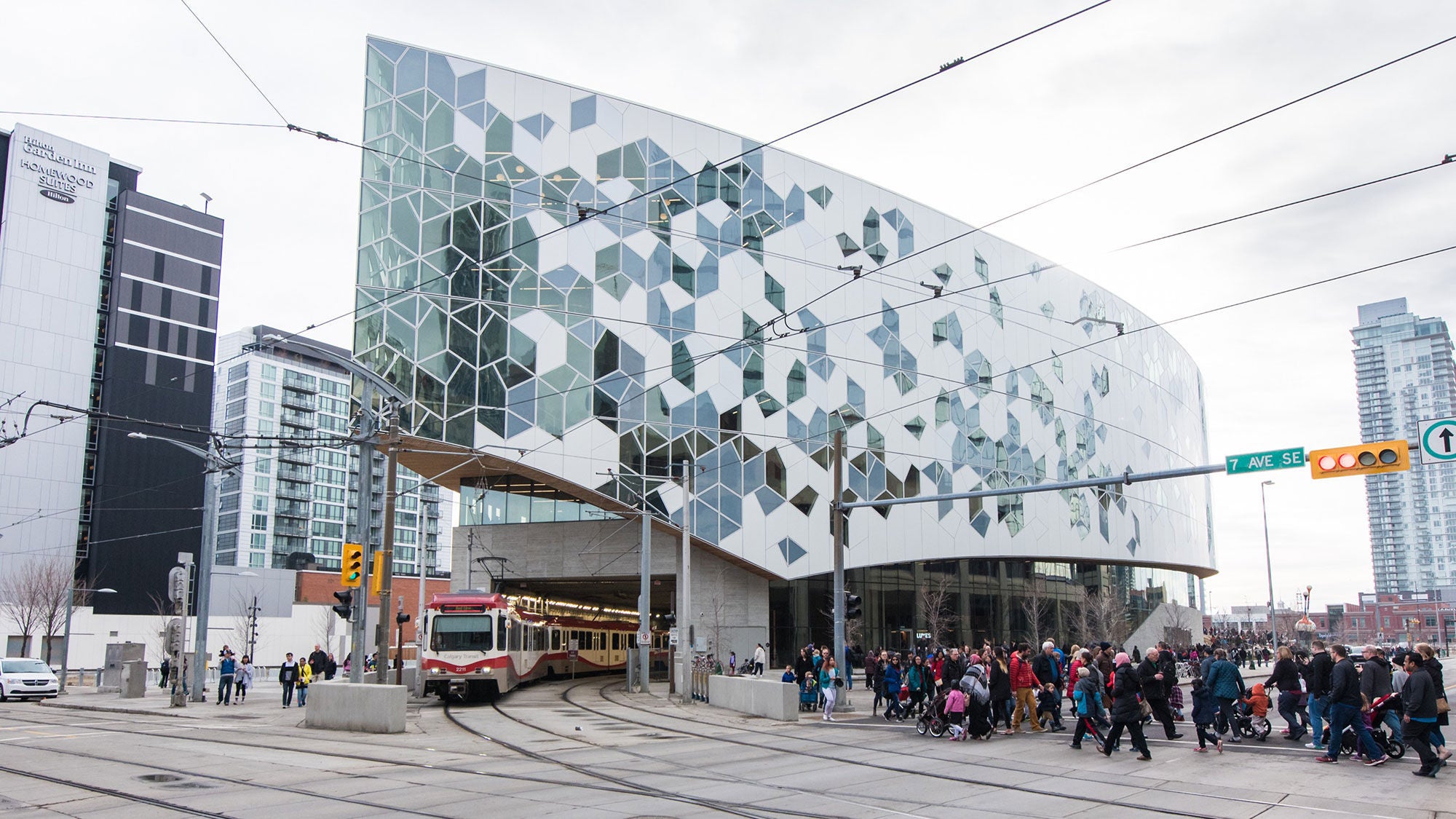 Exterior view of the Calgary Central Library featuring a red and white CTrain light rail vehicle emerging from a tunnel beneath the geometric glass building facade, with pedestrians crossing the street.