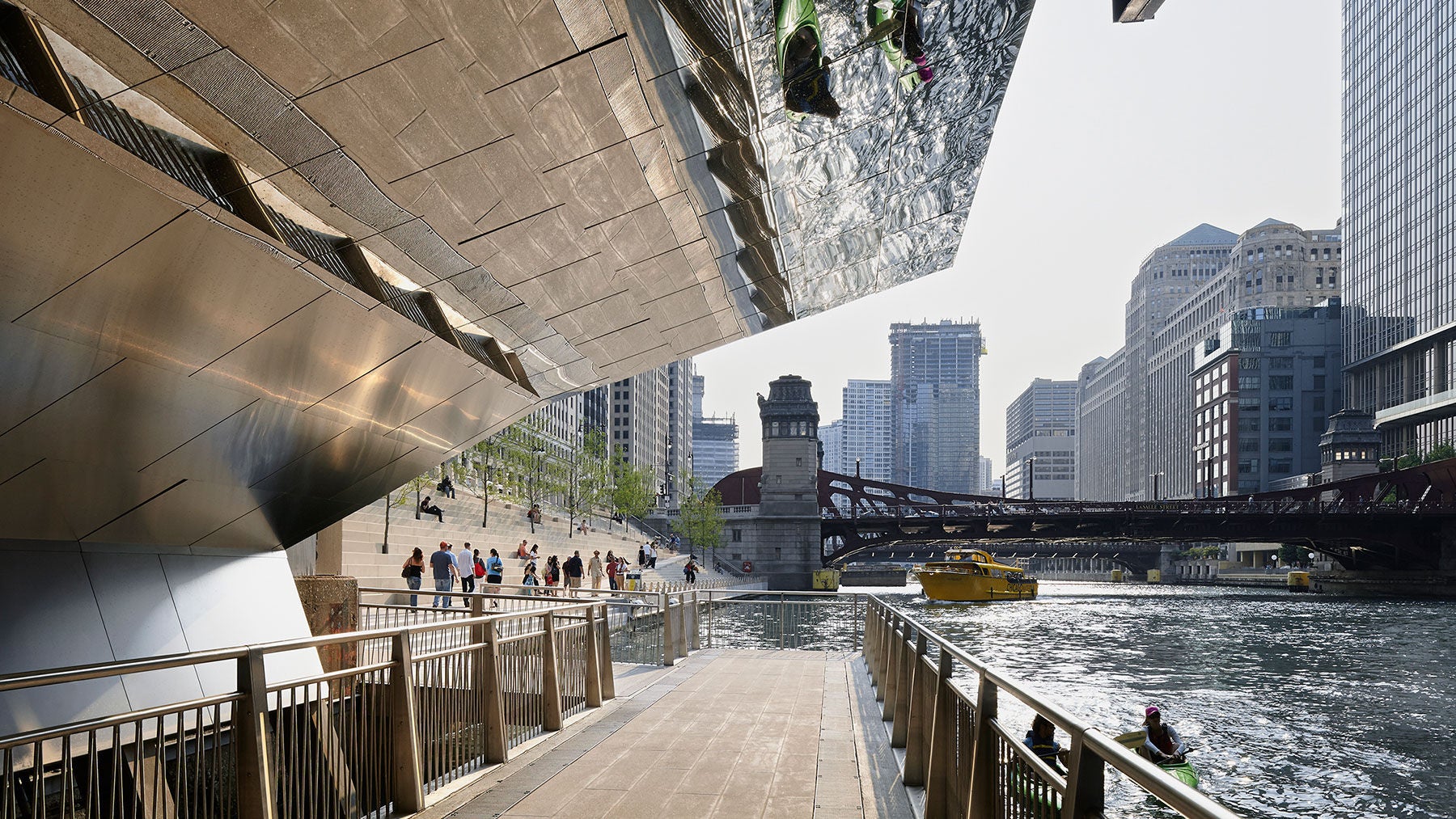 An image of the Chicago Riverwalk from under one of the bridges, showing the walking below the bridge and reflection of kayakers on the river in the metal surface above