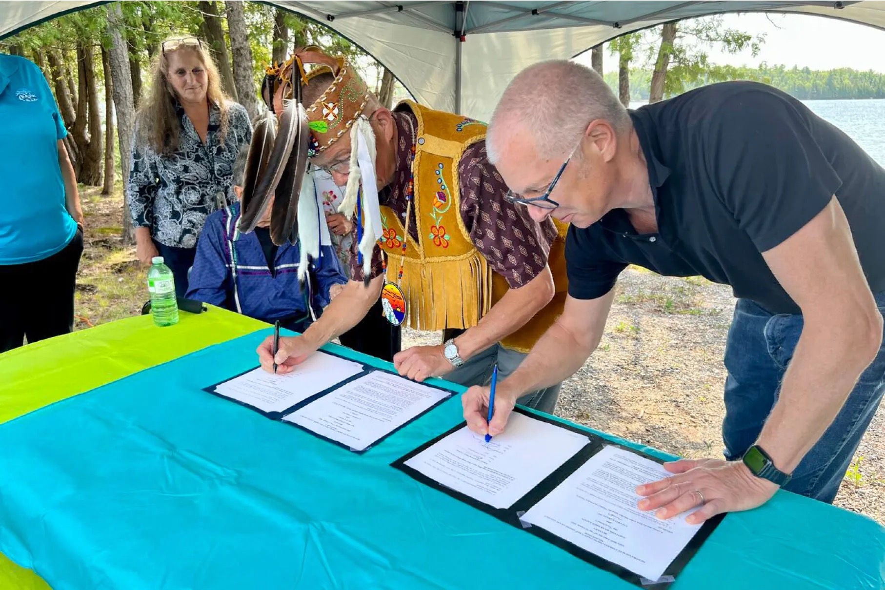 Deux hommes signent des documents; l’un est vêtu d’une tenue traditionnelle autochtone et l’autre est vêtu d’une tenue d’affaires décontractée