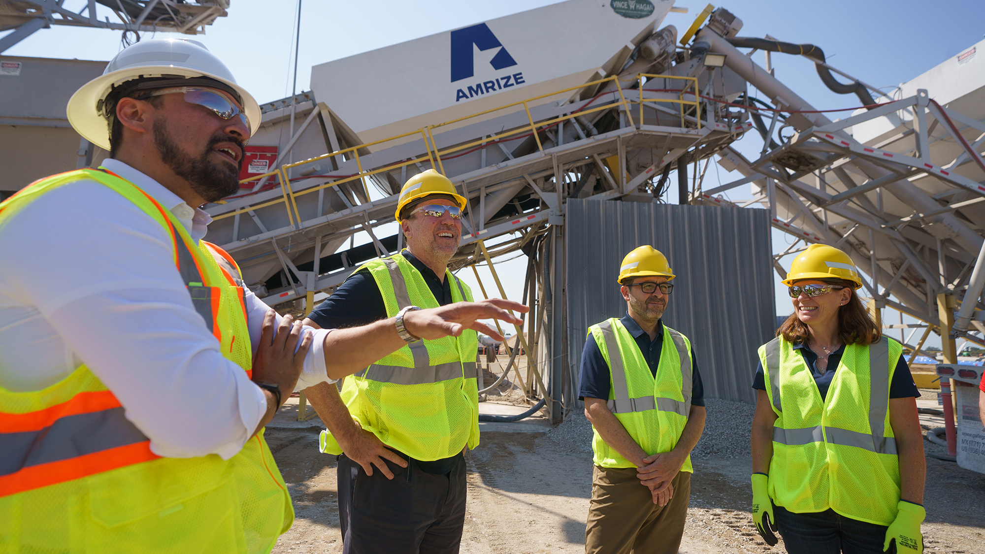 Four people in safety vests and hard hats stand in a construction area under bright sunlight, discussing near machinery labeled "AMRIZE," conveying teamwork and industry.