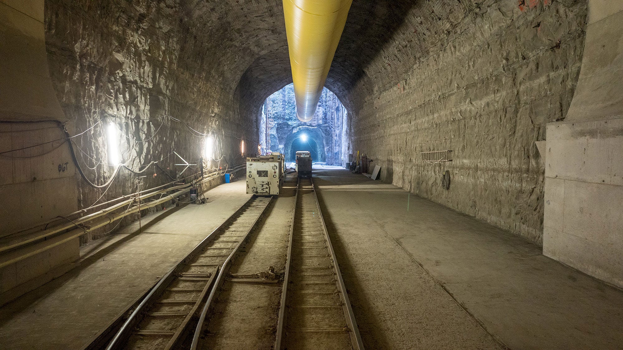 Underground tunnel construction site featuring temporary rail tracks, yellow ventilation ducting, and illuminated rock walls.