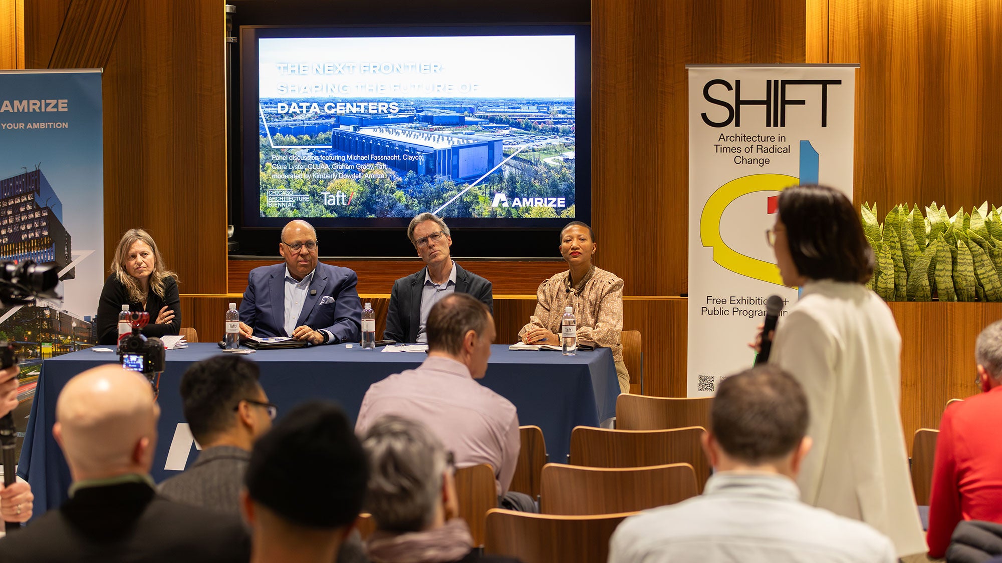 A group of three panelists sits at a table in front of a small crowd