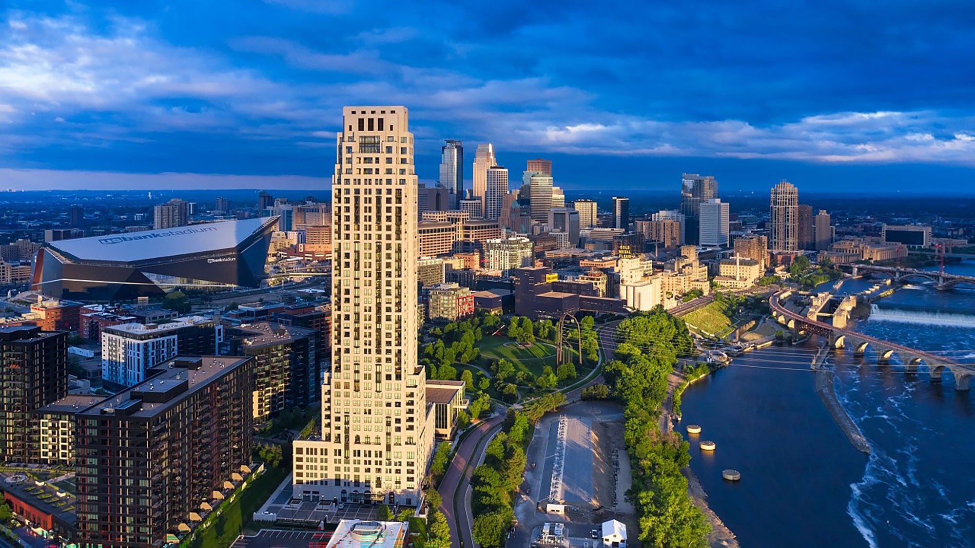 Aerial view of a cityscape at twilight, featuring ELEVEN, a tall building in the foreground, modern skyscrapers, a river with a bridge, and lush greenery.