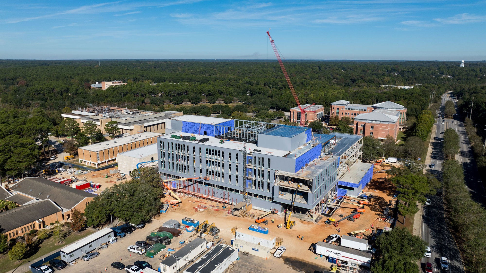 Aerial view of the USA's new Whiddon College of Medicine under construction with scaffolding and a crane, surrounded by trees and nearby structures, under a clear blue sky.