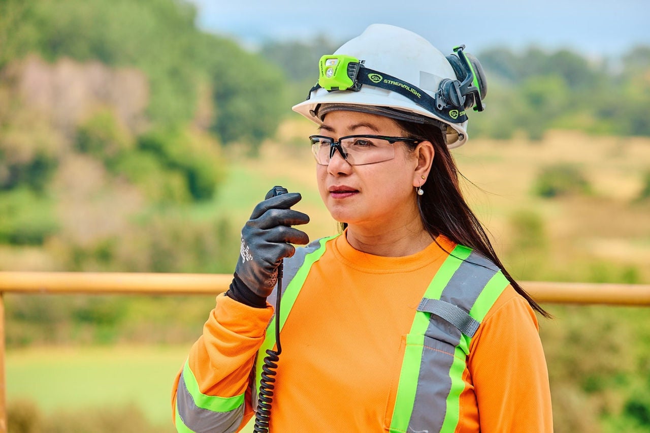 A female worker wearing safety vest, hard hat, and protective eyewear, speaking into a handheld radio at a construction worksite