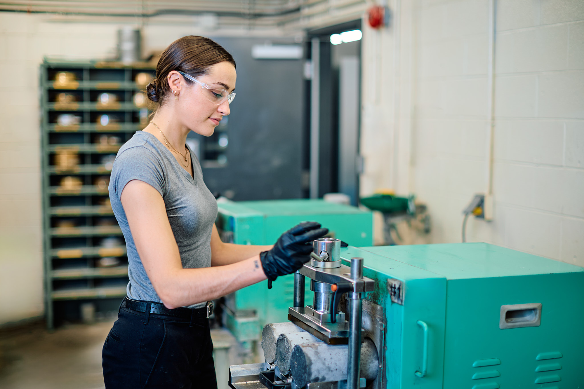 A woman in a  standing in a lab wearing gloves and protective glasses operating a machine.