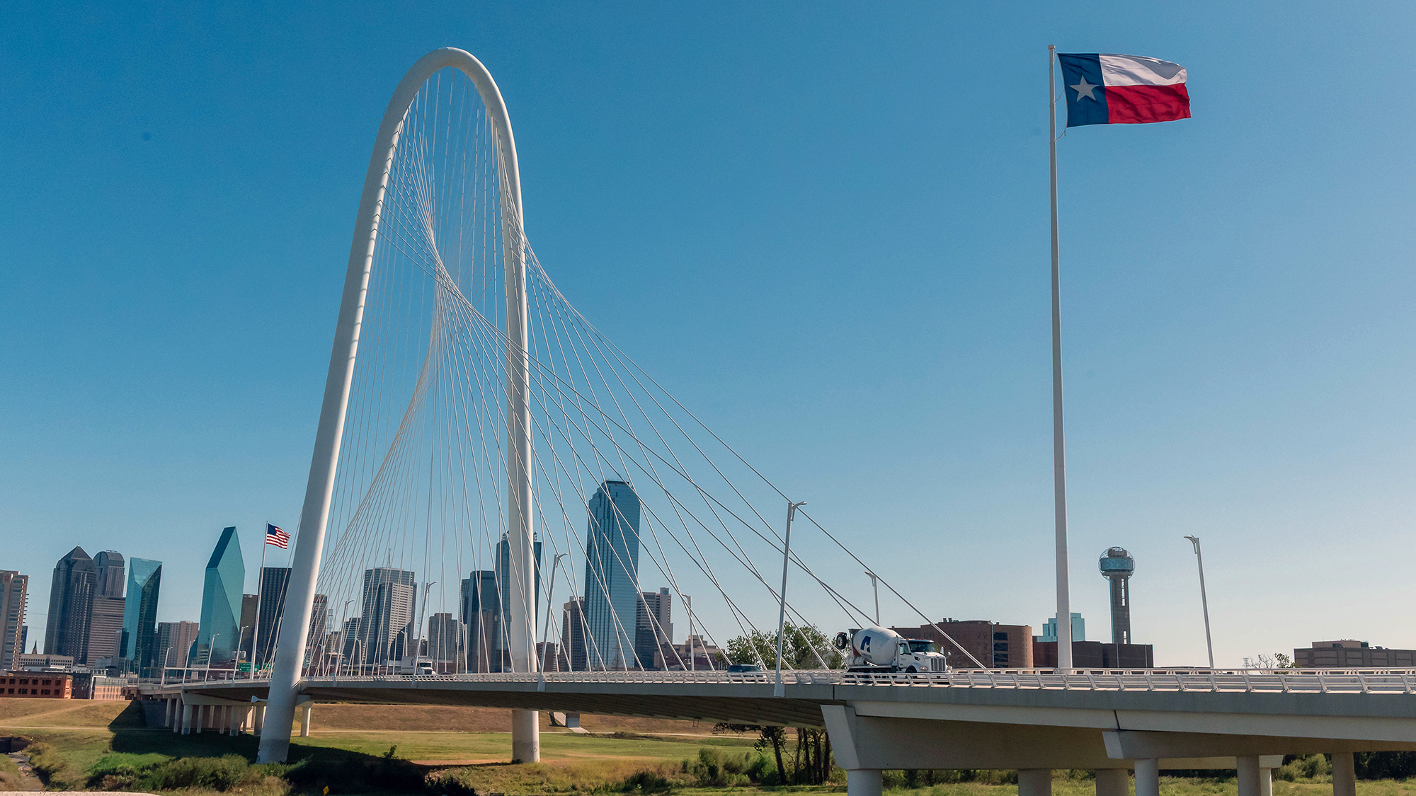 A bridge with a dramatic arch and cables, with a concrete truck passing over it and U.S. and Texas flags to the side