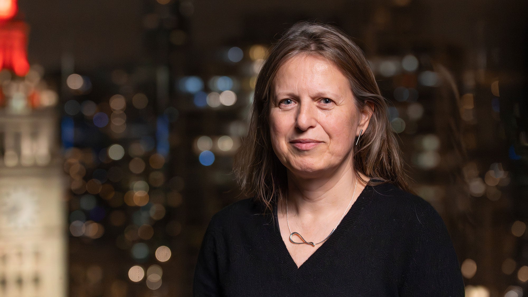 A female architect and professor looks into the camera, as lights from the city of Chicago twinkle behind.
