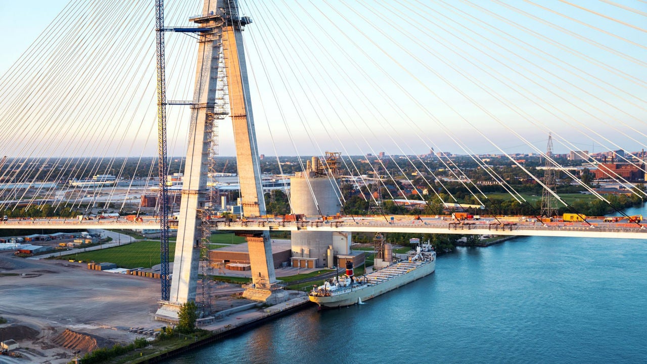 Gordie Howe International Bridge tower and deck construction with freighter ship docked along the Detroit River.
