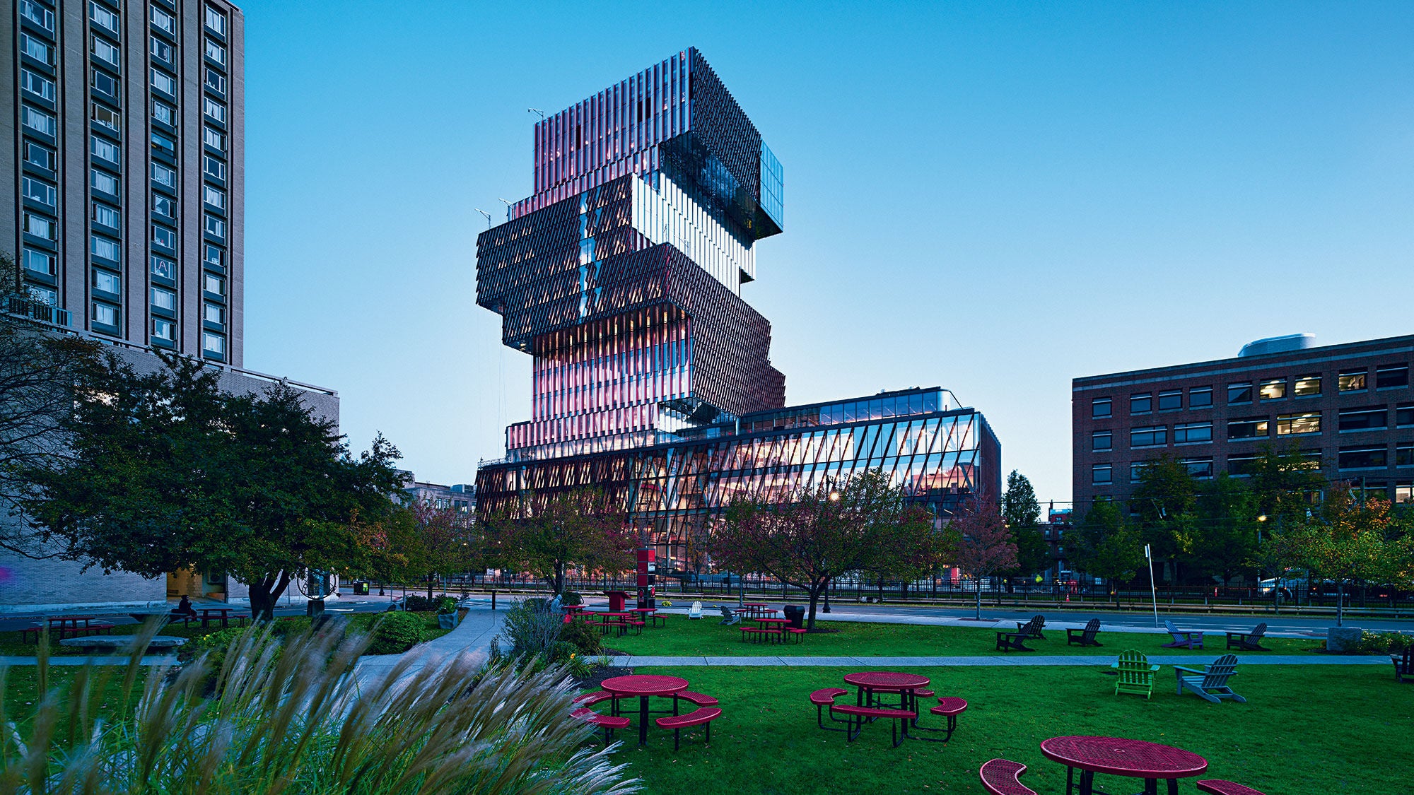 A tall building made up of block shapes rises into a blue sky behind a green city park