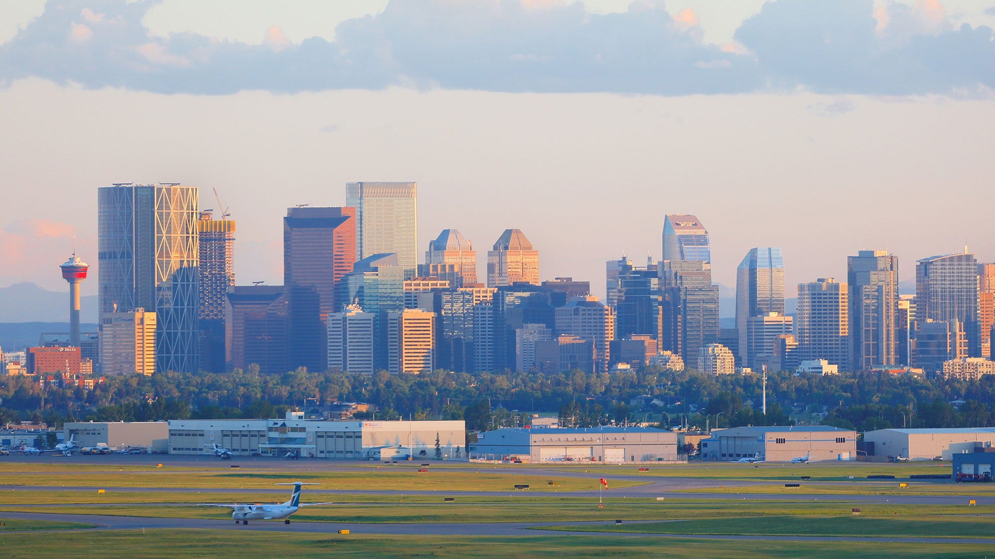 A view of a WestJet plane on the Calgary International Airport runway with the downtown skyline, illuminated by sunset light in the background.