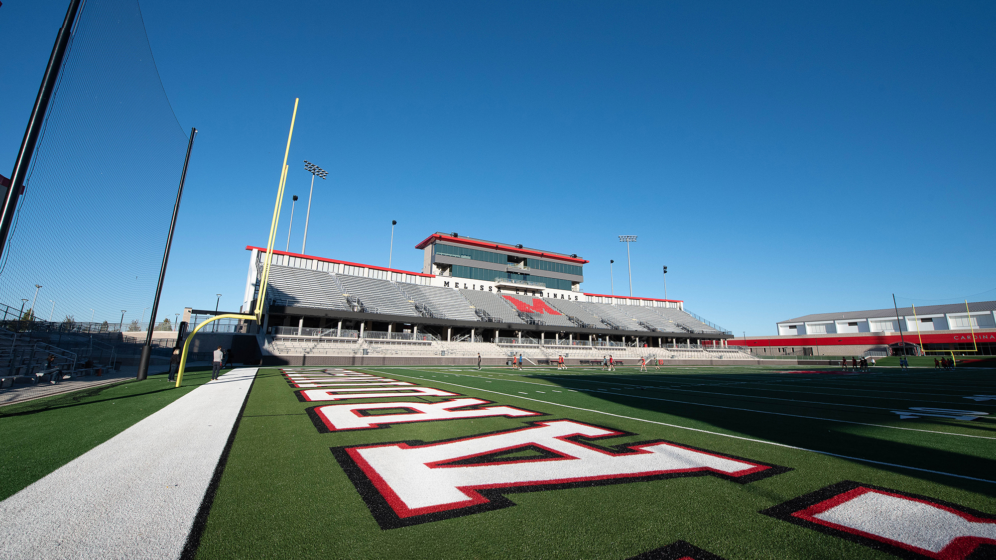 Wide-angle view of an empty football stadium with green turf and large red and white letters in the foreground. Bleachers flank both sides under a clear blue sky.