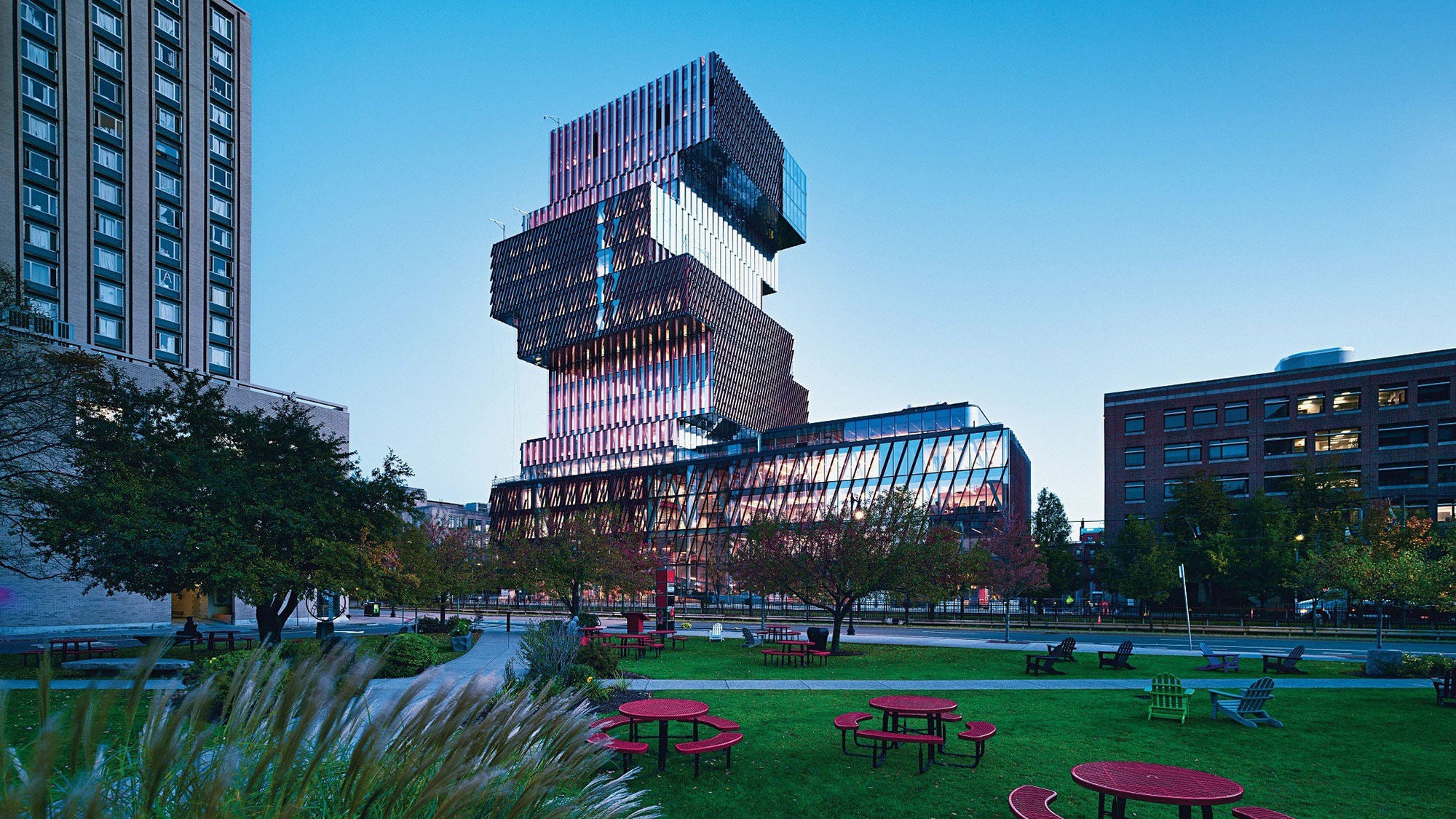 Modern building at Boston University with stacked, reflective glass sections at dusk. Surrounded by a green park with red picnic tables and trees.