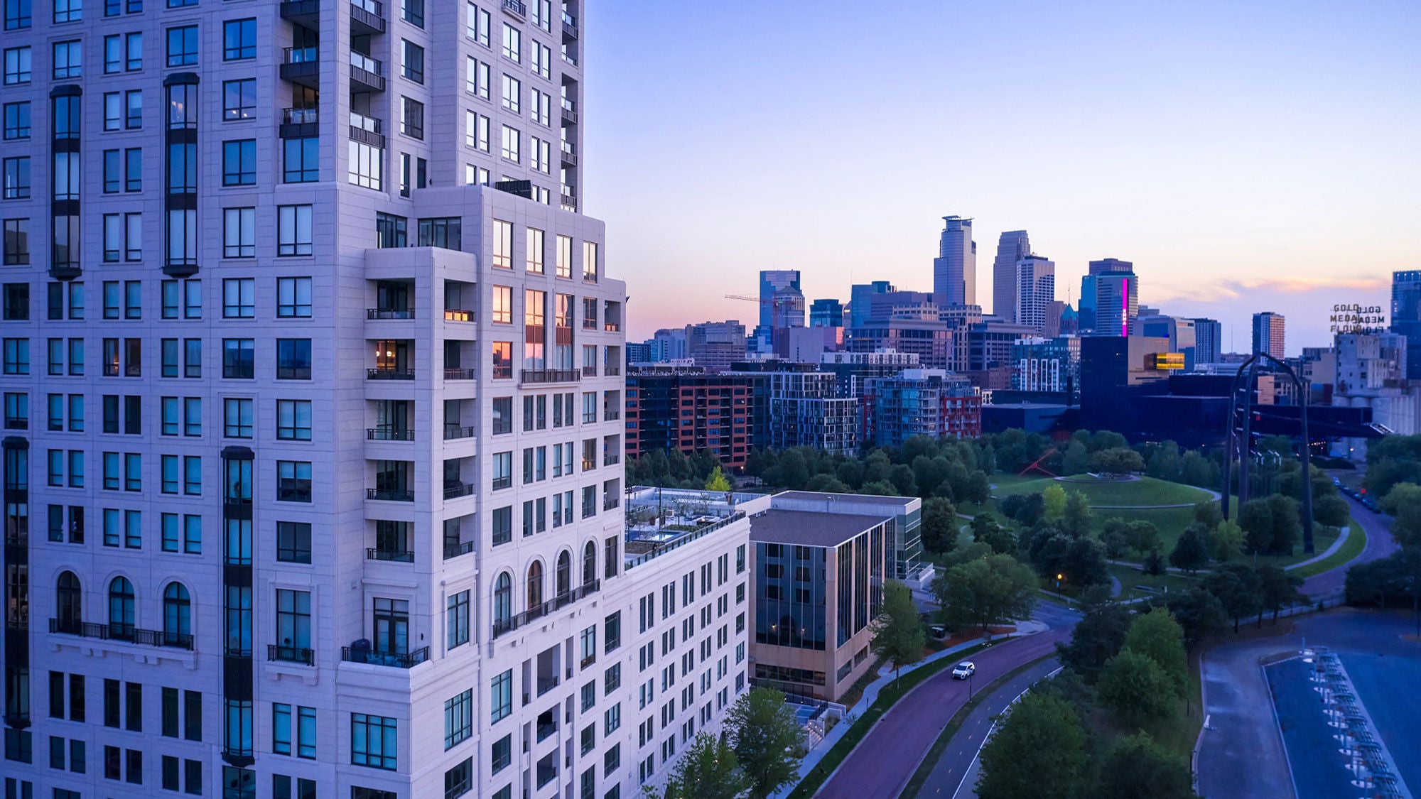Cityscape at dusk featuring ELEVEN, a modern high-rise in the foreground with lit windows. The skyline includes various buildings and greenery under a soft pastel sky.