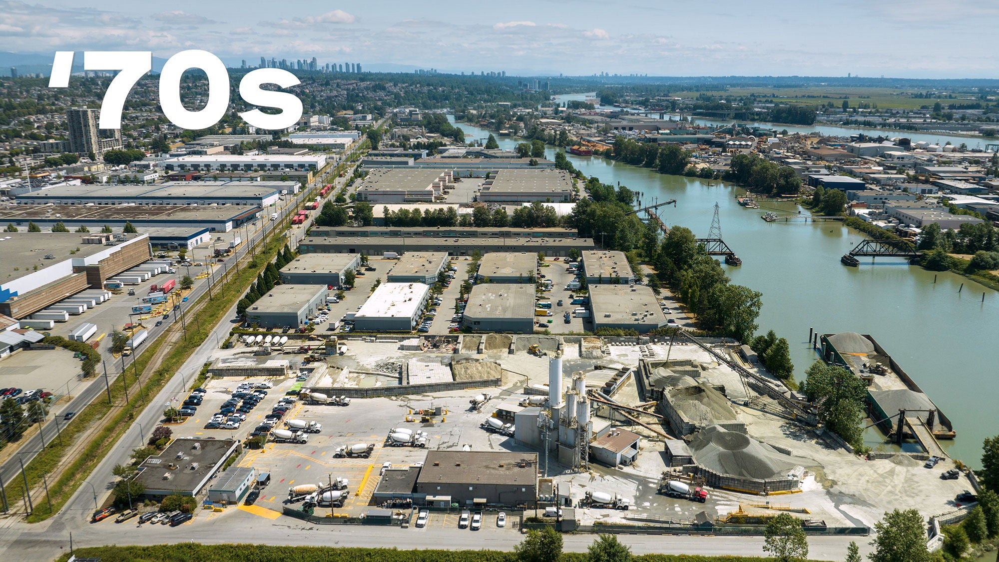 Vue aérienne d’une zone industrielle avec plusieurs grands entrepôts et une rivière. La silhouette urbaine et le paysage urbain lointain sont visibles sous un ciel bleu clair. Inscription « années 70 » en caractères gras blancs au-dessus.