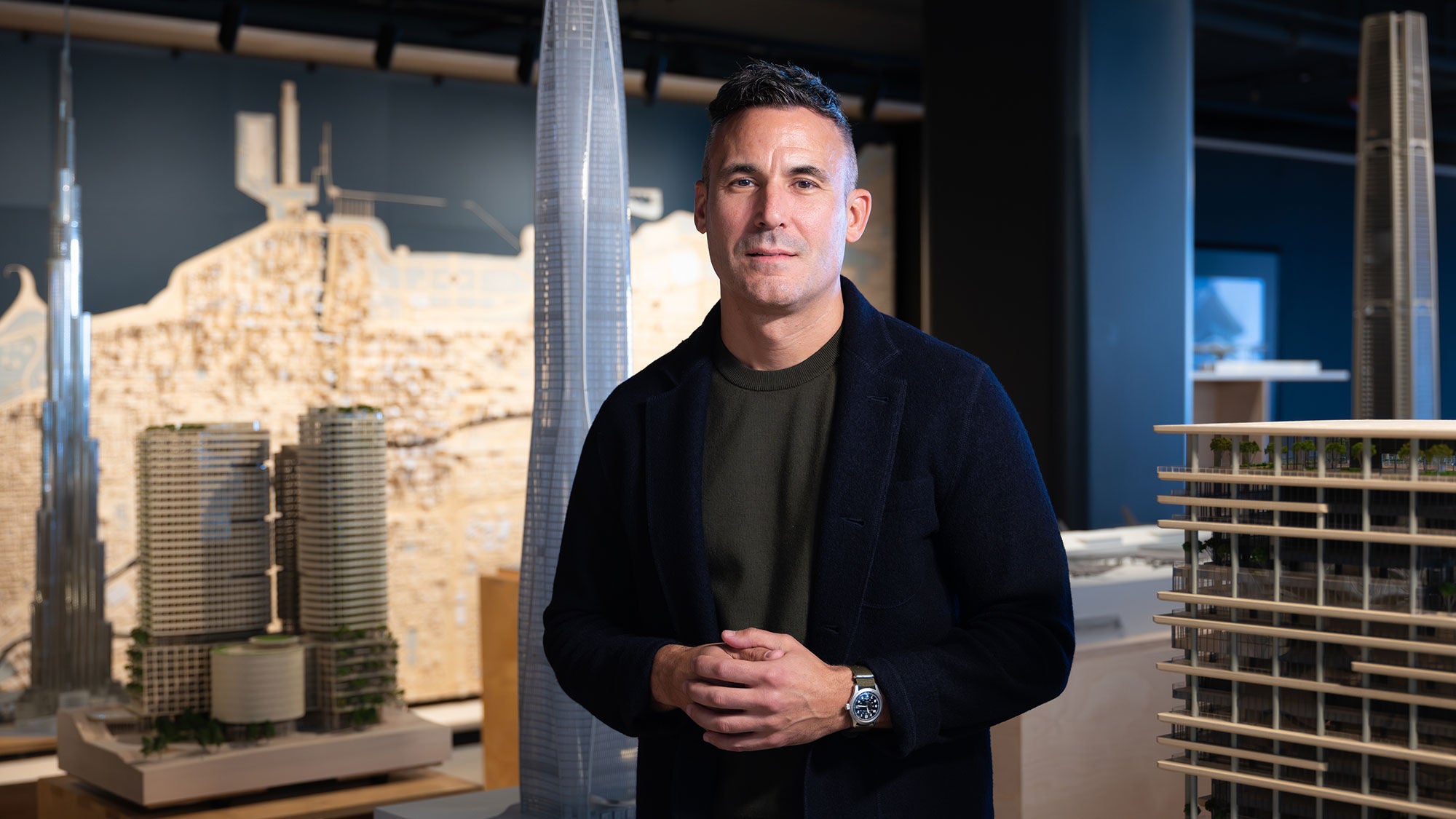 An architect stands next to an architectural model in a studio. He looks into the camera with his hands folded in front of him.