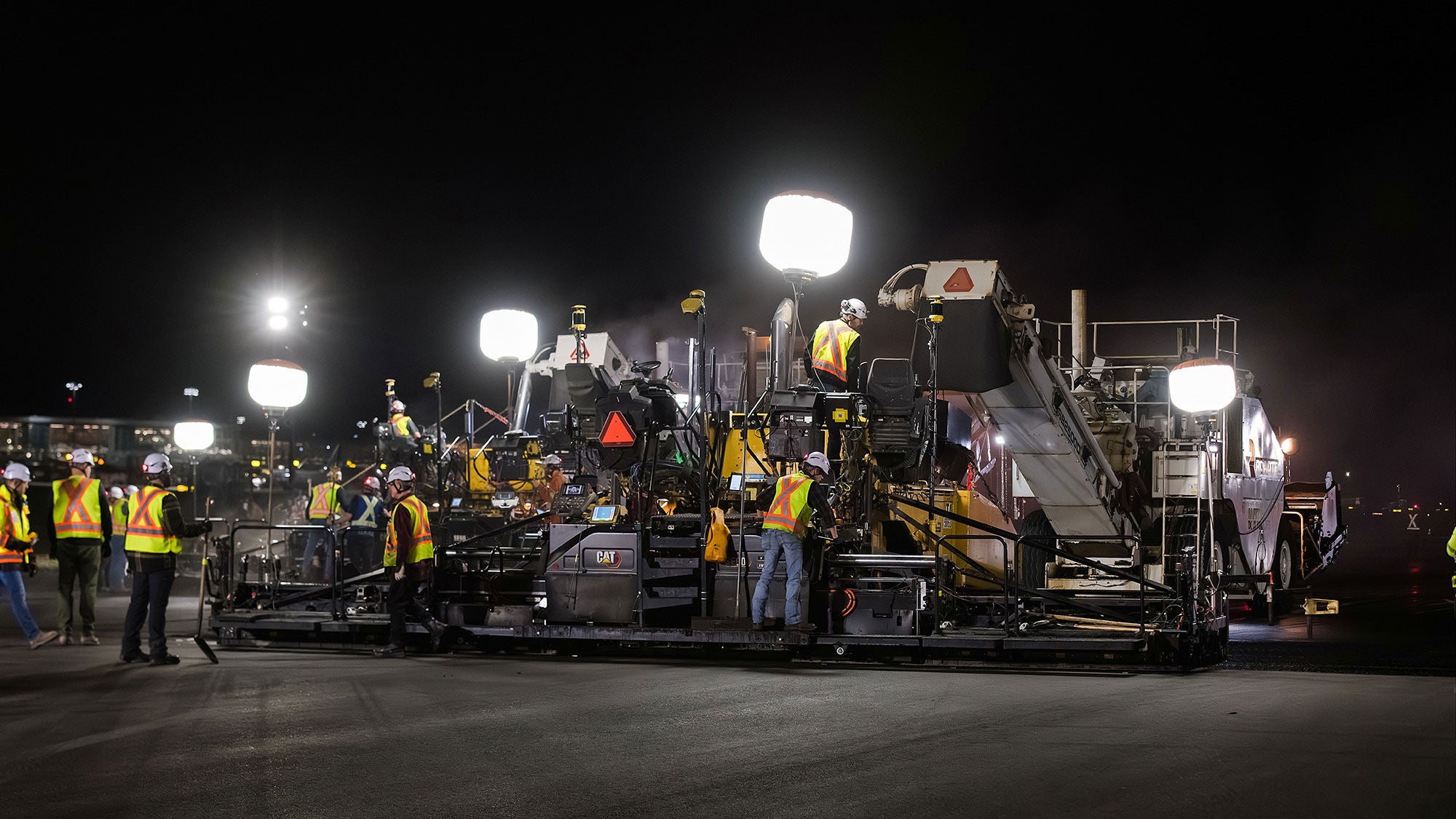 Night roadwork scene with workers in reflective vests operating machinery under bright floodlights. The atmosphere is focused and busy.