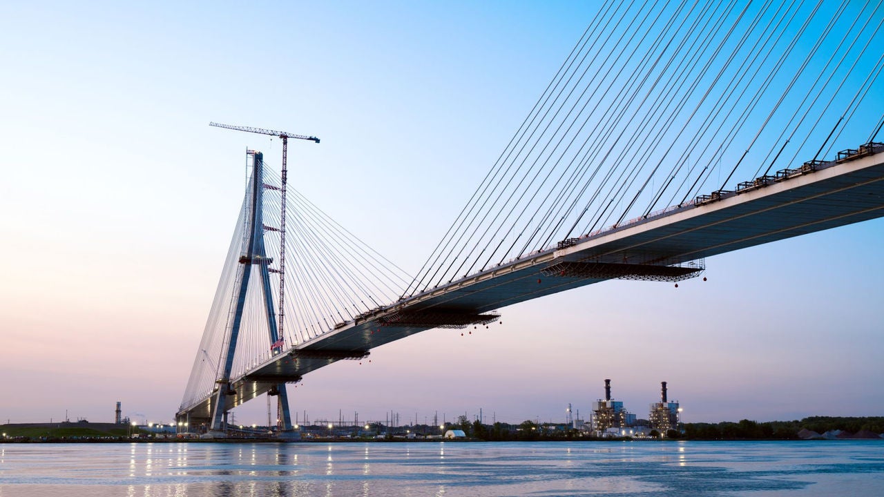 Cable-stayed deck and tower of the Gordie Howe International Bridge under construction at dusk with glowing city lights along the water.