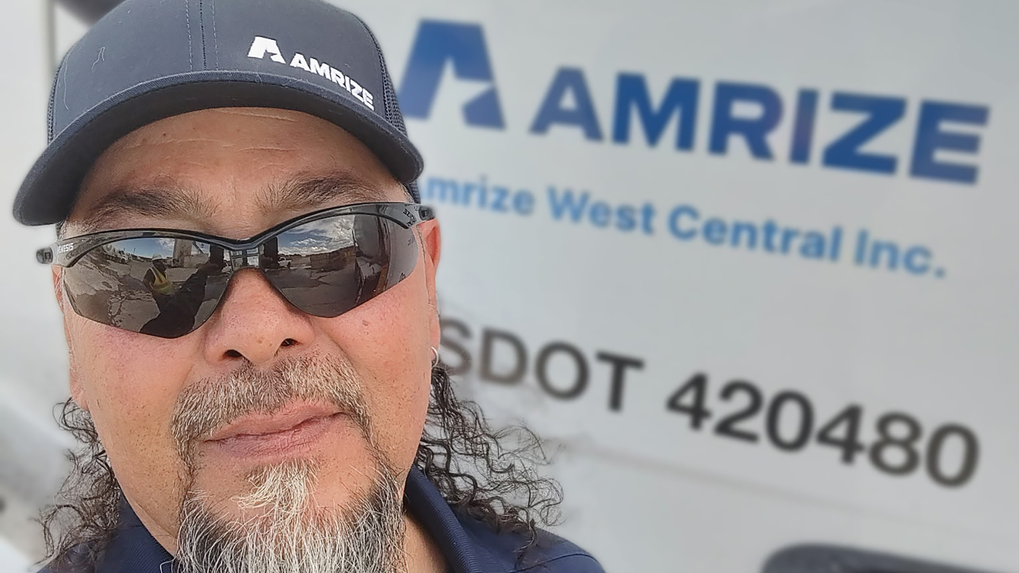 A man in sunglasses and a baseball hat standing in front of a truck with the name “Amrize” on the side
