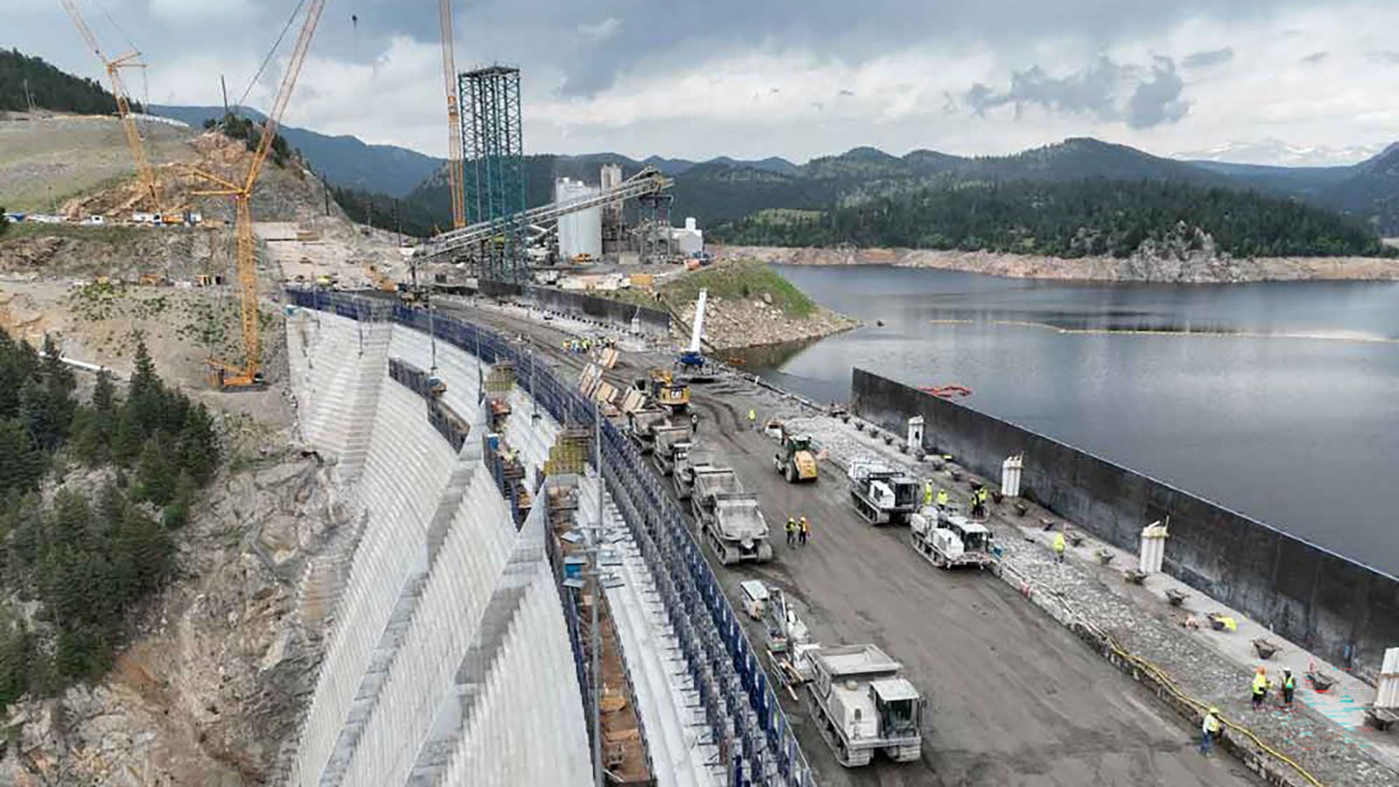 Aerial view of a dam construction site with cranes and machinery on a cloudy day. The partially built dam curves along a rocky hillside near a large reservoir.