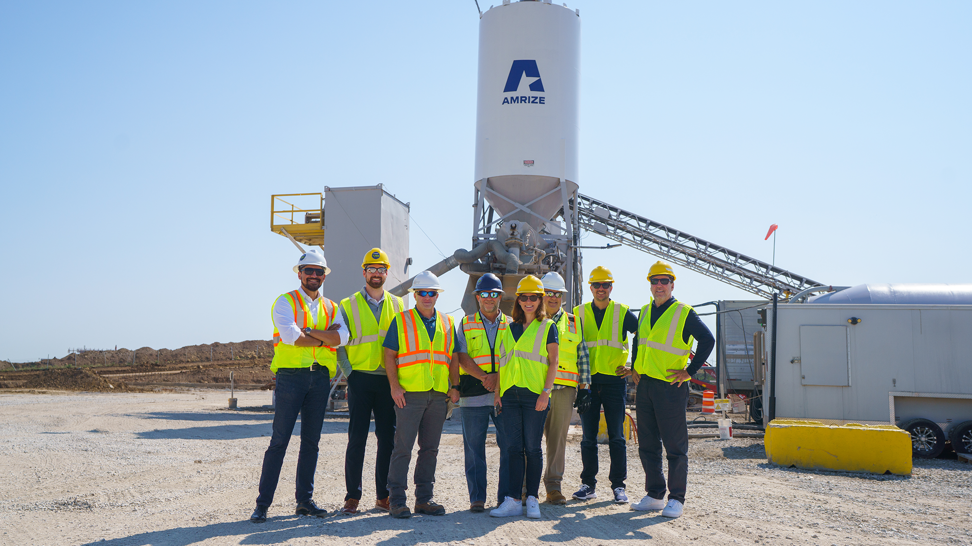 A group of eight people wearing bright safety vests and hard hats smiling on a sunny day at a construction site with equipment in the background.