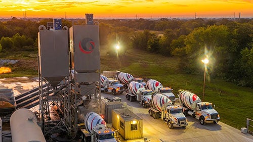 A collection of concrete mixing trucks lined up below a silo at sunset
