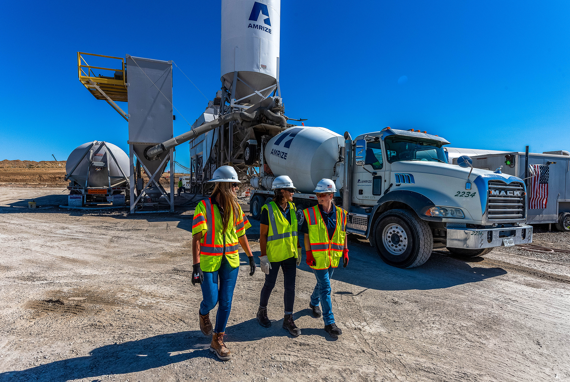 Three women walking down a dirt road wearing protective gear in front of a cement truck