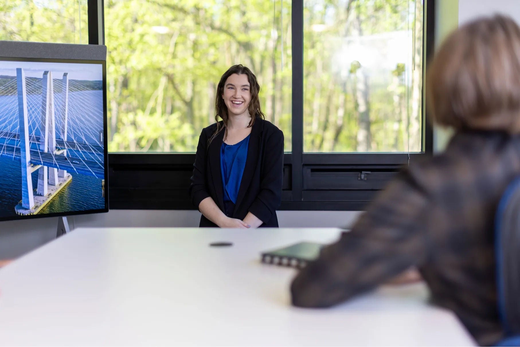 A woman stands in front of a room beside a screen with the image of a bridge on it