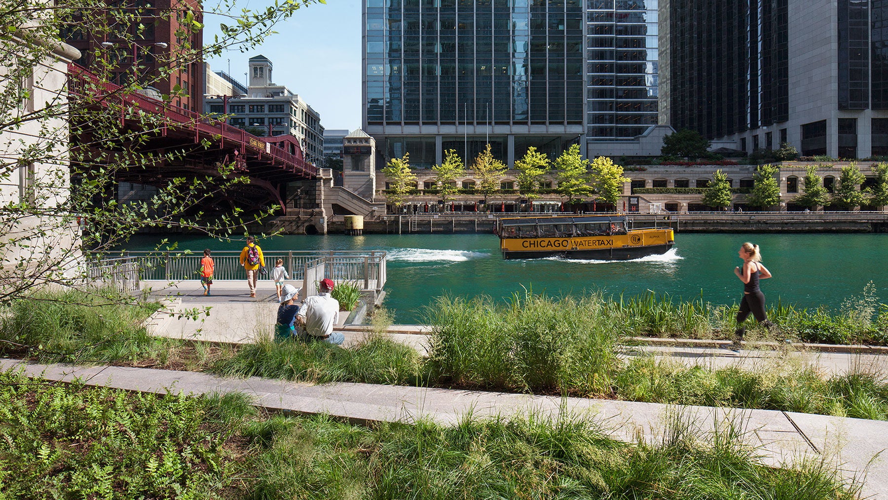 A runner jogs along the Chicago Riverwalk. Behind her you can see the river with a water taxi passing by. 