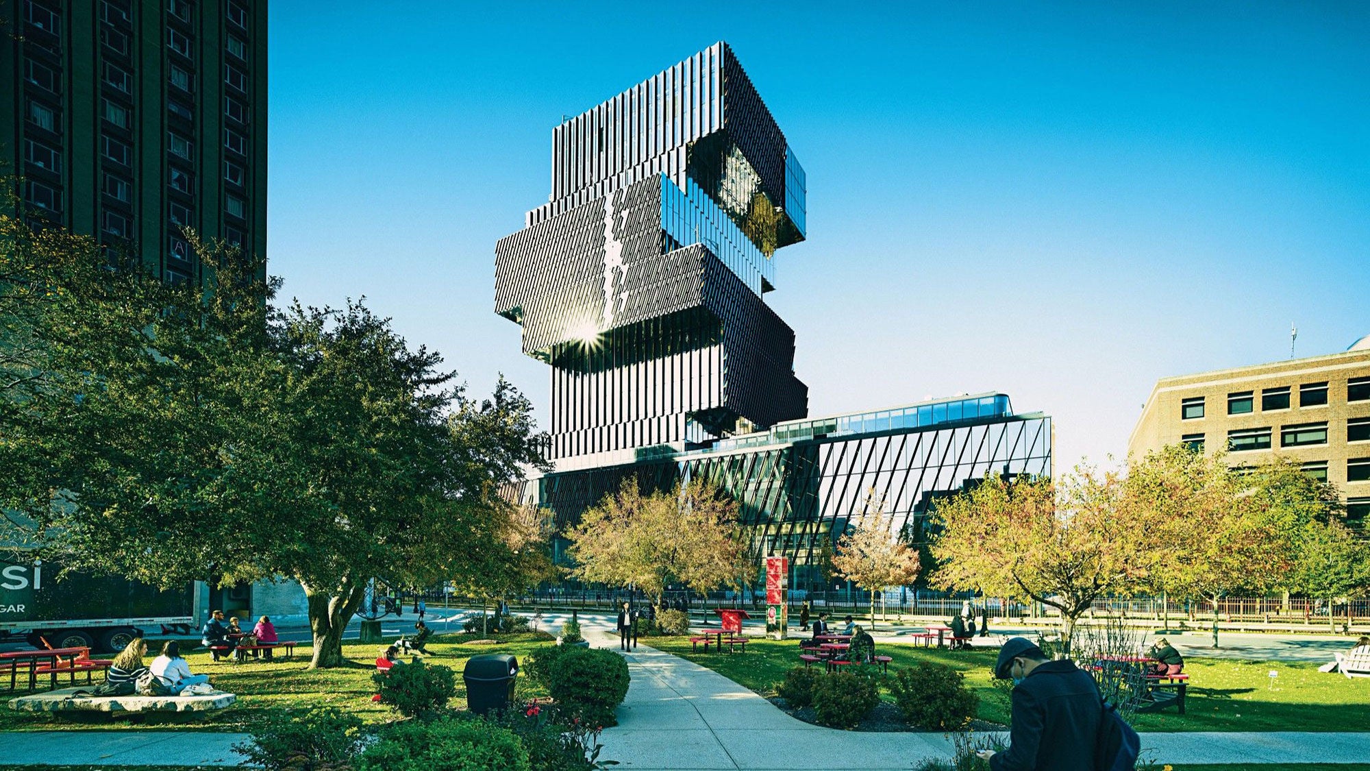 A modern, geometric building at Boston University with stacked, offset sections and reflective windows towers over a green park filled with trees, benches and people relaxing.