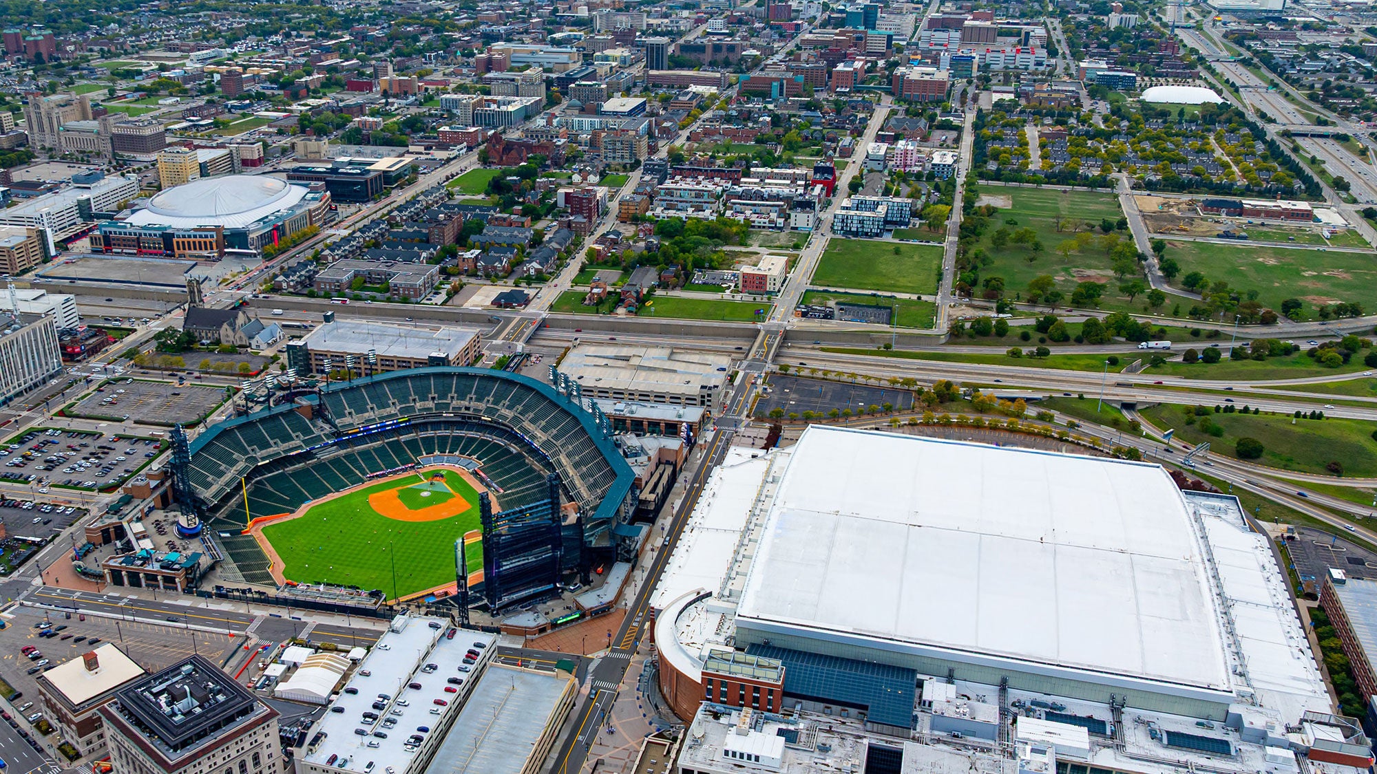 High aerial drone photograph of downtown Detroit featuring Comerica Park baseball stadium and the white roof of Ford Field side-by-side.