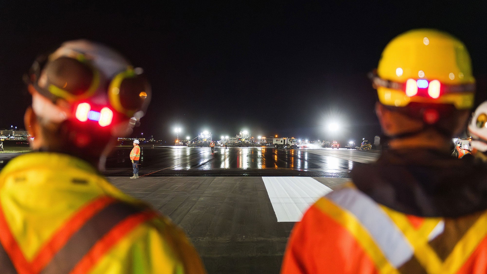 Two construction workers in safety gear and hard hats observe a brightly lit tarmac at night, creating an atmosphere of focus and diligence.
