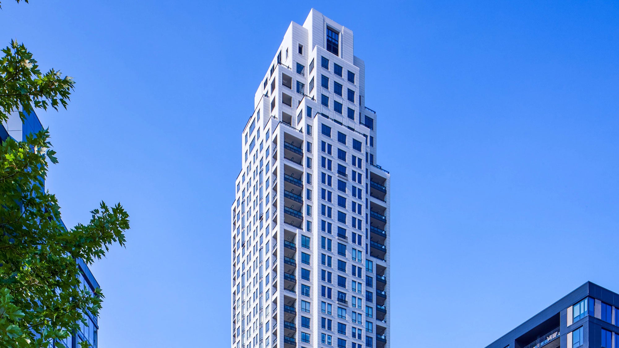 Tall modern skyscraper, ELEVEN, with a geometric design against a clear blue sky, flanked by trees on the left and another building edge on the right.