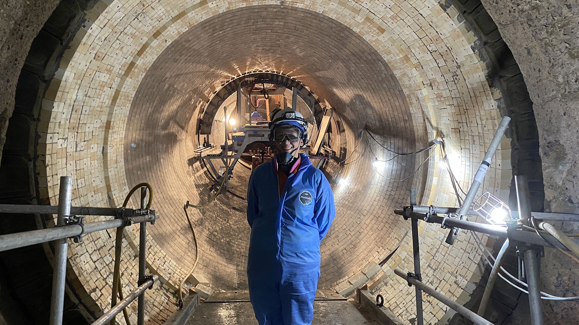 A woman wearing blue overalls and safety gear stands in front of a tunnel.