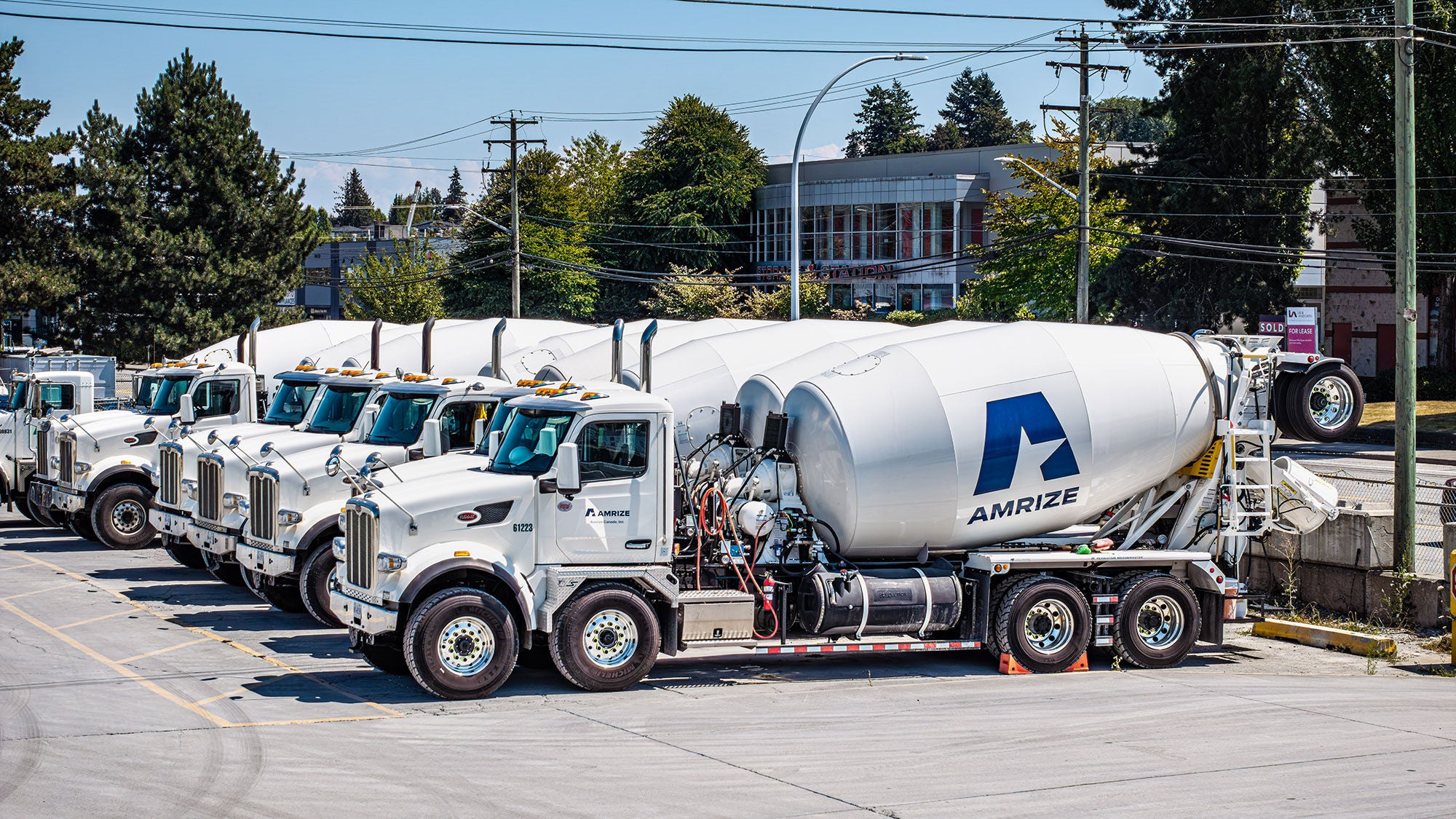 A row of white ready-mix concrete trucks with "AMRIZE" branding parked in an industrial lot. Trees and buildings are visible in the background under a clear sky.