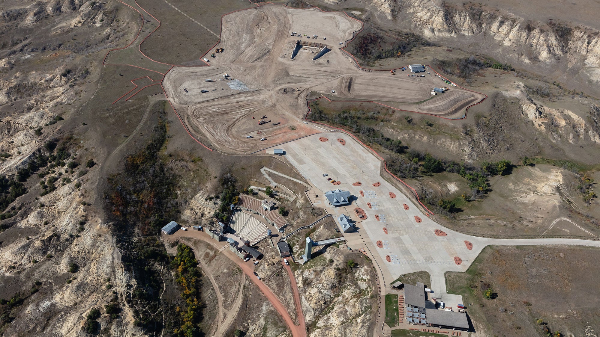 High-angle aerial view of the Theodore Roosevelt Presidential Library construction site in Medora, North Dakota, showing early-stage earthworks, foundation grading, and heavy machinery on a plateau overlooking the Badlands.