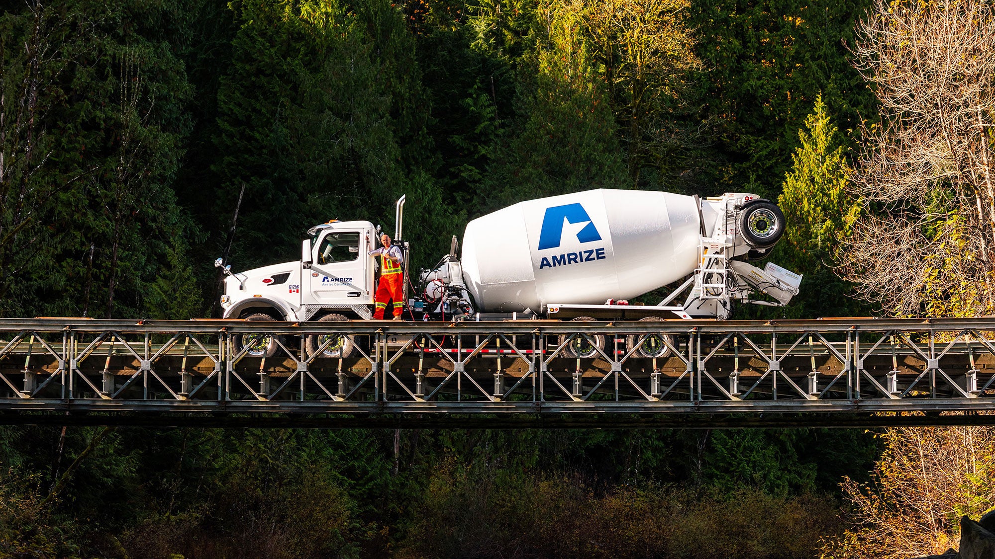 A forestry green backdrop featuring a white Amrize truck centered in the middle on a bridge crossing with a driver waving in front of the RMX truck.