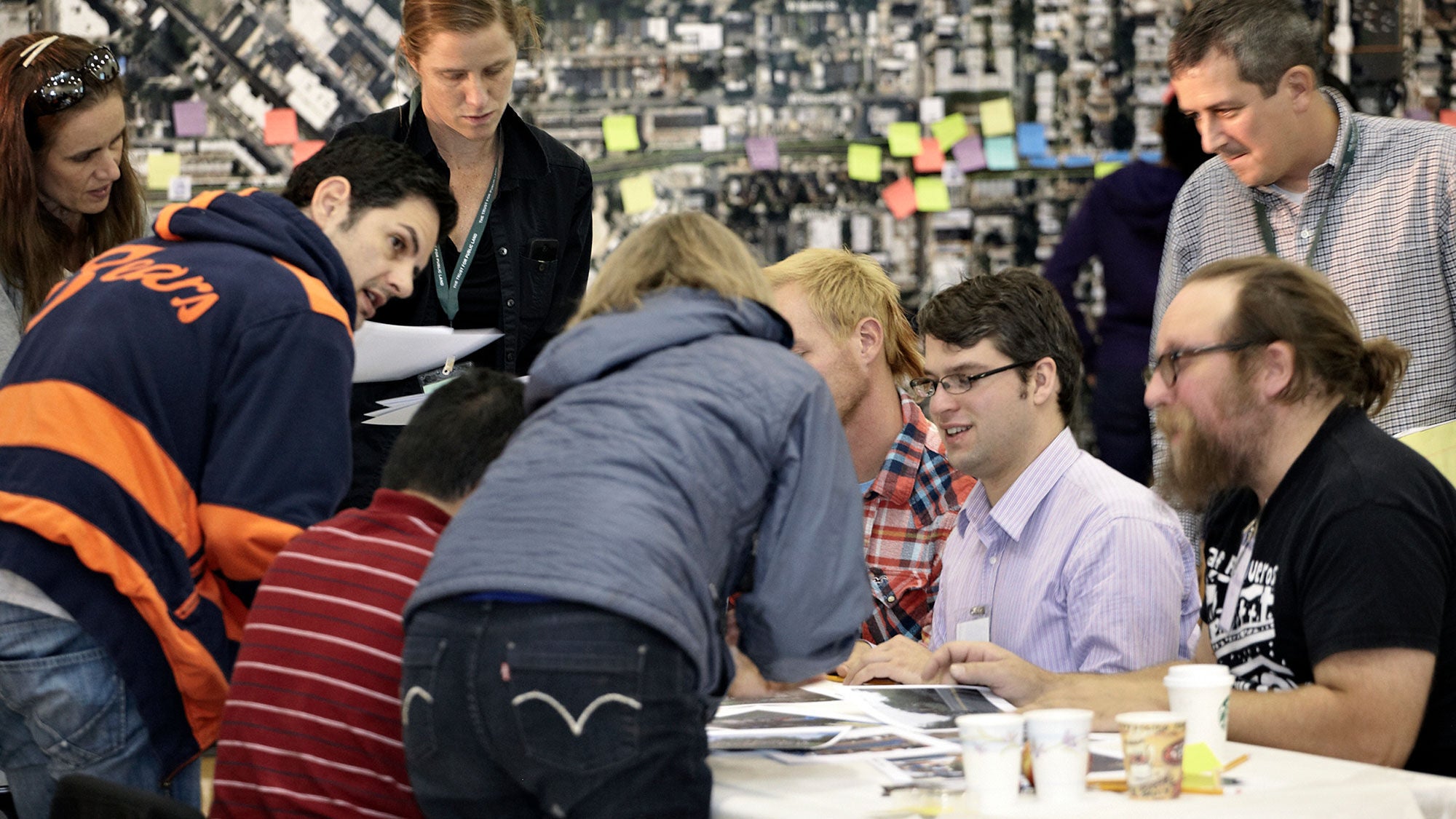 A group of people gather around photos on a table and hold a discussion. There is a large map with sticky notes on it behind them.