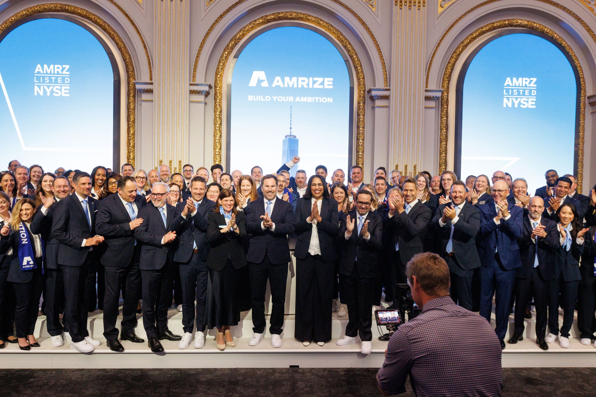 A group of people stand on stage clapping while a photographer takes pictures in the foreground
