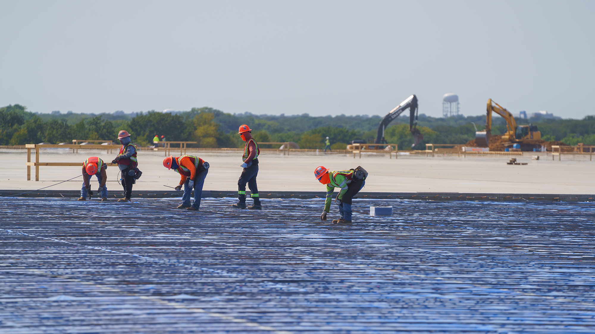 Construction workers in orange hard hats and vests apply coating to a large flat surface. Excavators and trees are in the background under a clear sky.