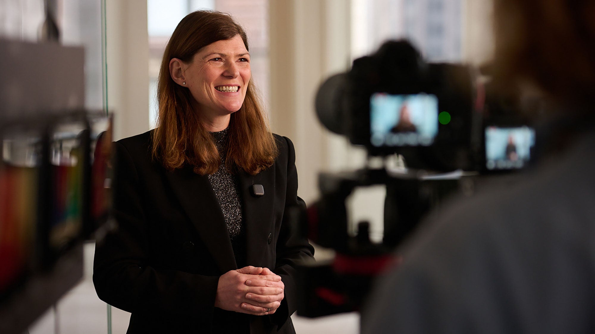 A woman in a suit smiles as a camera captures the moment in the foreground.