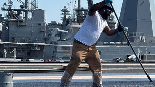 A man in a white t shirt spreads sealant on the deck of a large navy vessel
