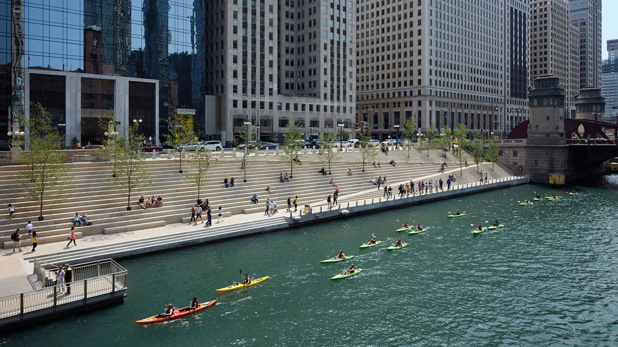 A fleet of kayakers paddle up the Chicago River while people walk past or sit on the Riverwalk benches.