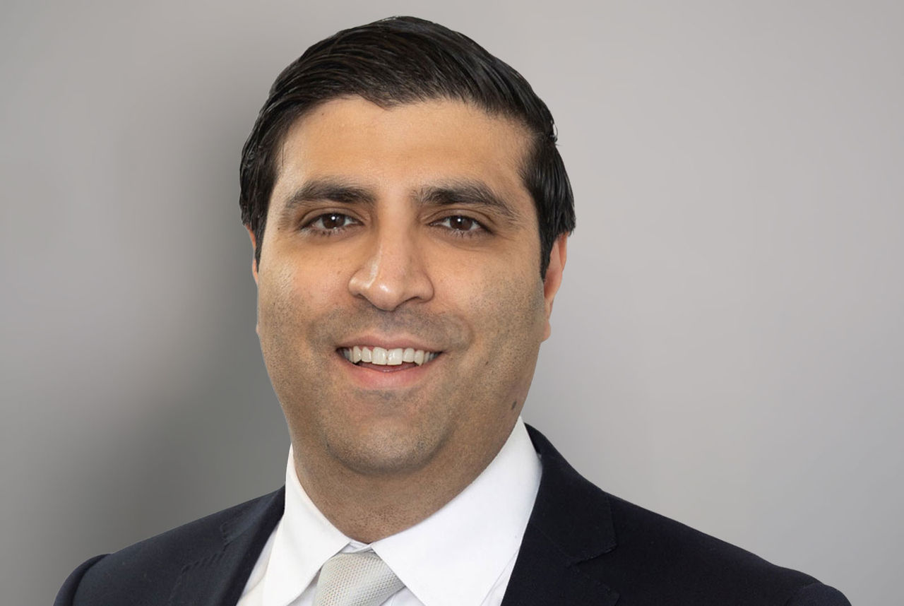 Professional headshot of Aroon Amarnani, wearing a black blazer and white collared shirt against a neutral gray background.